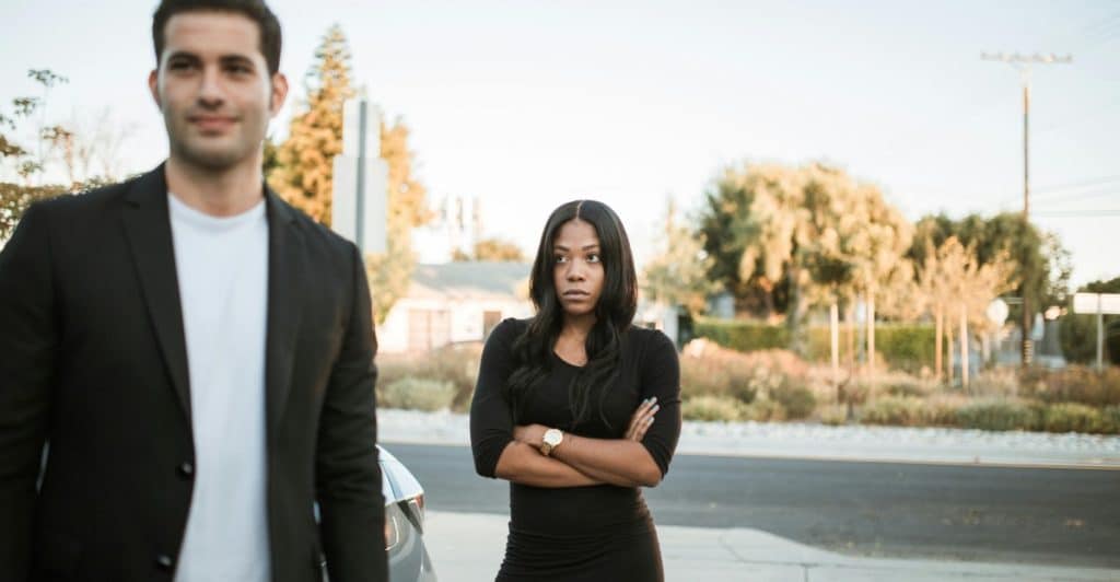 A man in a suit looking off to the side while a woman with crossed arms looks in a different direction outdoors.