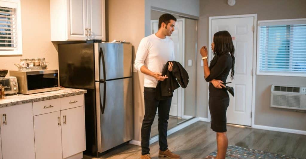 A man and a woman are standing in a kitchen, having a conversation. The man is holding a jacket.