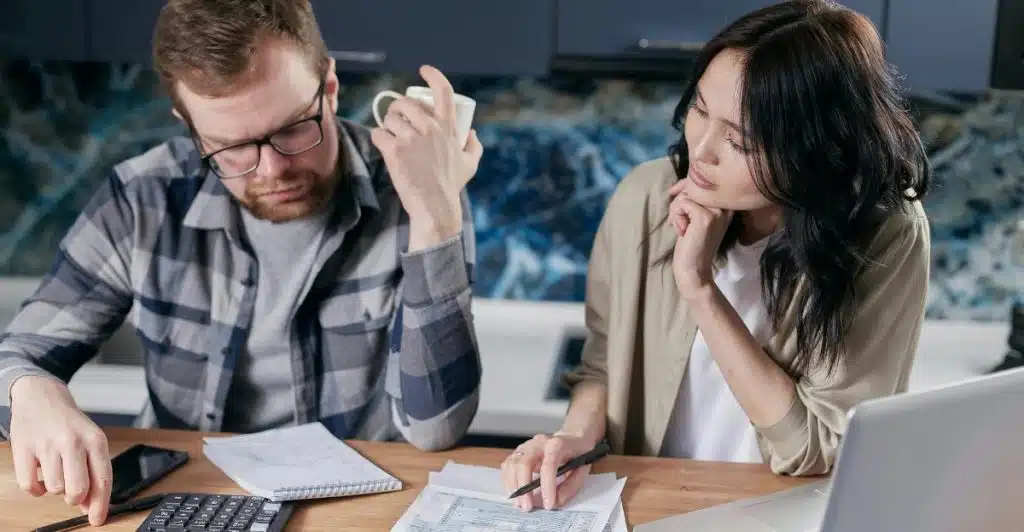 A Man and a woman sitting at a table with a laptop, a calculator and some documents.