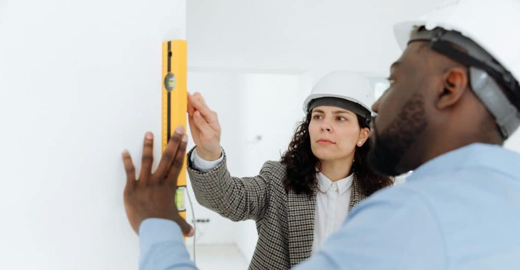 A man and a woman wearing white hard hats, holding a yellow spirit level against a white wall, working on a construction project.