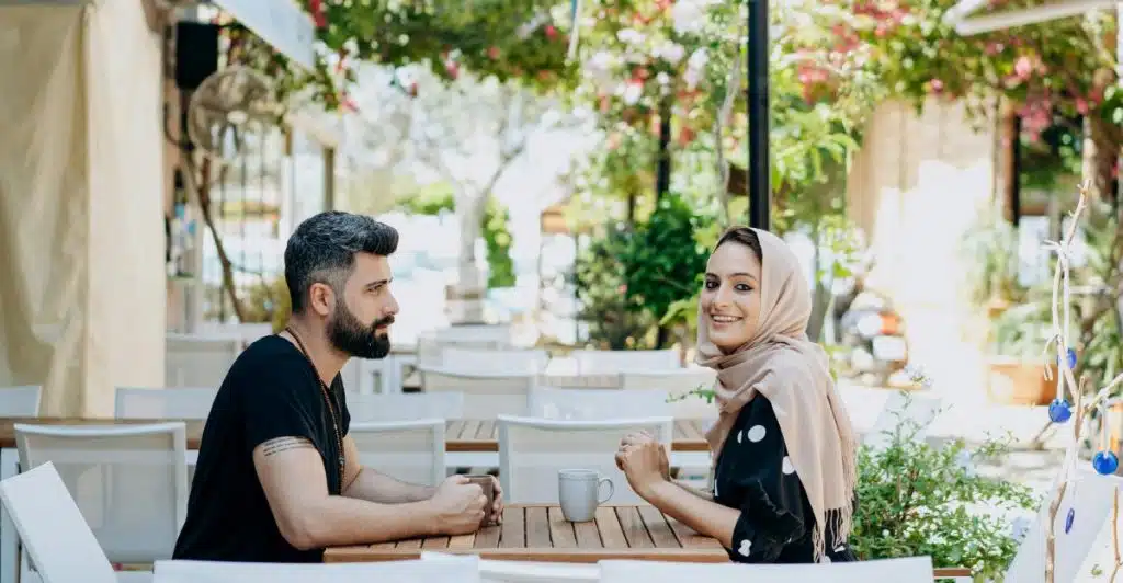 A man and a woman sit facing each other at an outdoor cafe table with cups, looking at each other and talking.
