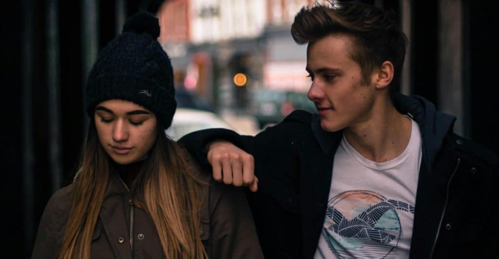 A young man looking at a young woman with his arm on her shoulder in a dimly lit tunnel.