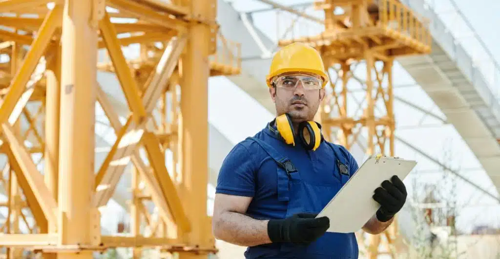 A man in a yellow hard hat, safety glasses, and ear protection, wearing blue overalls and gloves, holds a clipboard at a construction site with yellow metal structures in the background.