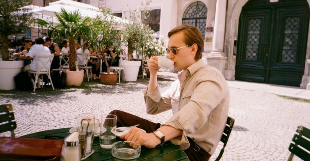 A man in a light brown shirt and sunglasses sitting at a green outdoor table with a coffee cup in his hand.