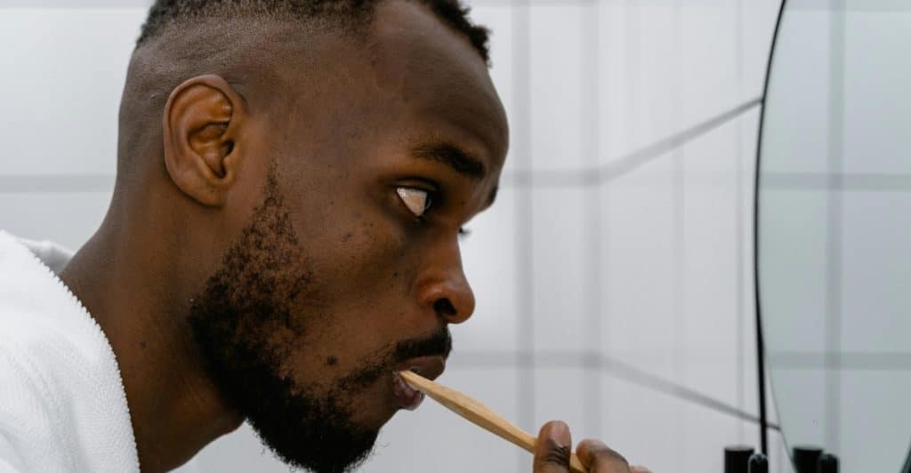 A man in a white robe is brushing his teeth with a wooden toothbrush in a bathroom