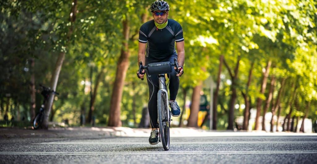 A man wearing cycling gear riding a bicycle on an asphalt path lined with green trees.