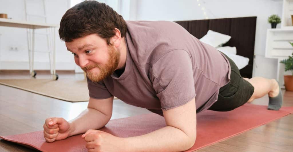 A man with brown hair and a beard doing a plank exercise on a red yoga mat in a room.