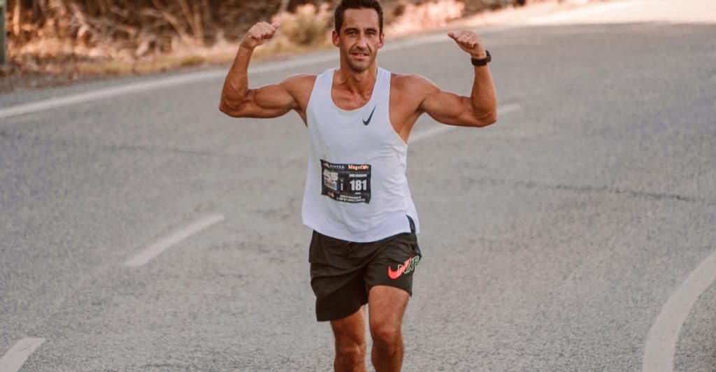 A muscular man wearing a white tank top and running shorts flexing his biceps while running on an empty asphalt road.