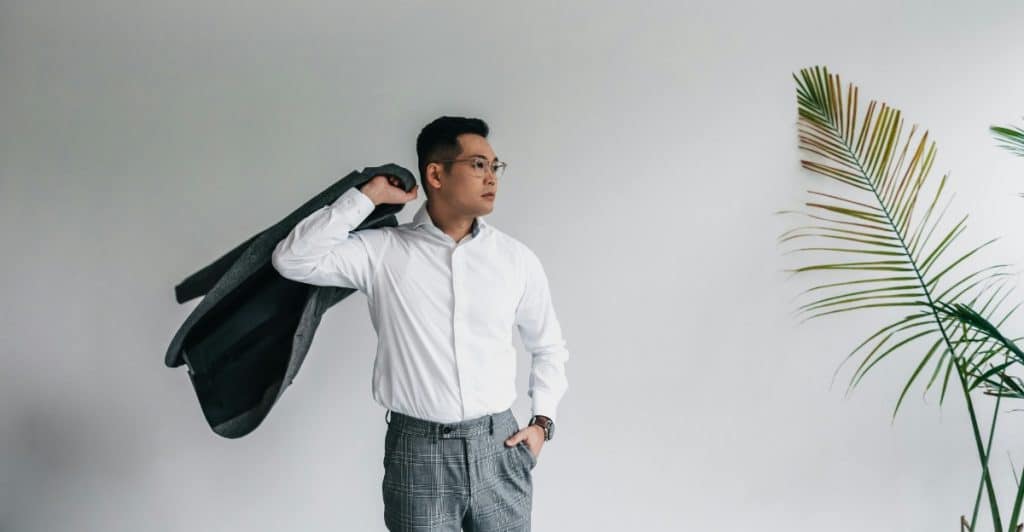 A man in a white dress shirt and grey plaid pants is standing against a white wall, holding a dark grey jacket over his shoulder, wearing glasses and looking to the side with a plant is to his right.
