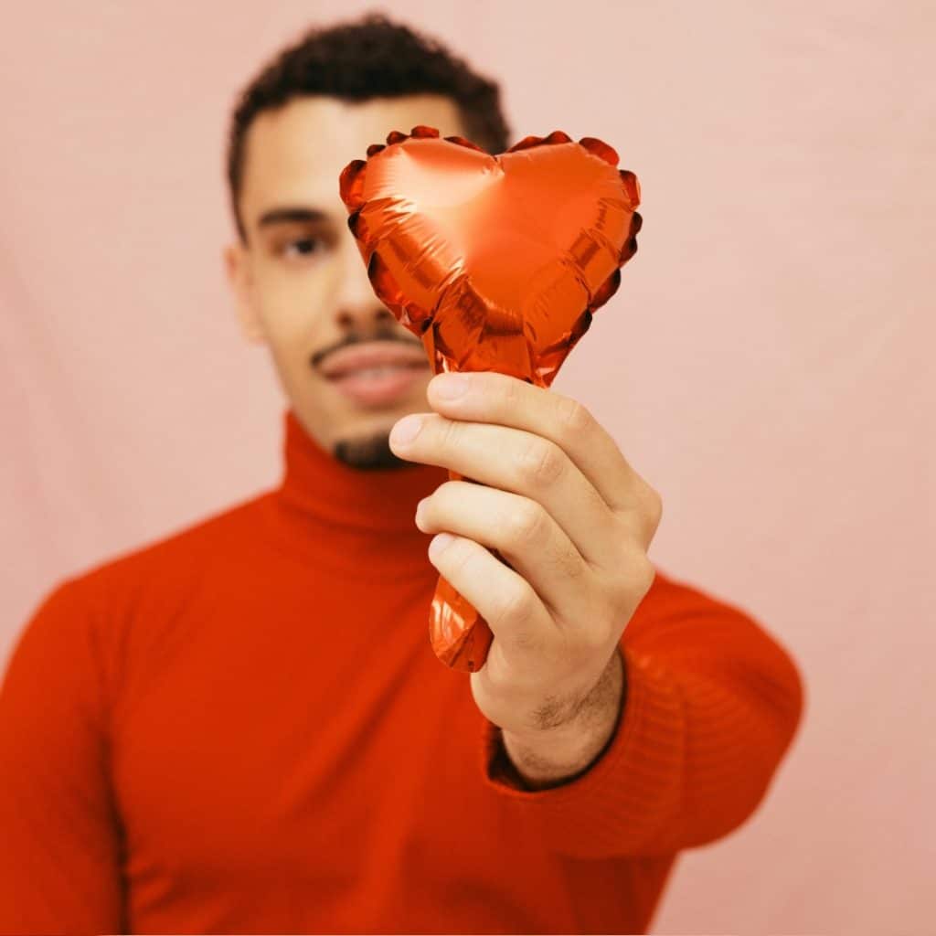 A man with short dark hair and a mustache, wearing a red turtleneck sweater, holding a red heart-shaped balloon against a pink draped background.