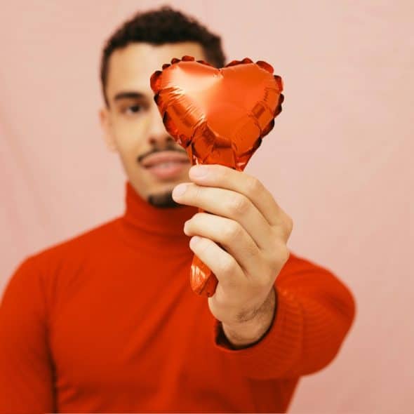 A man with short dark hair and a mustache, wearing a red turtleneck sweater, holding a red heart-shaped balloon against a pink draped background.