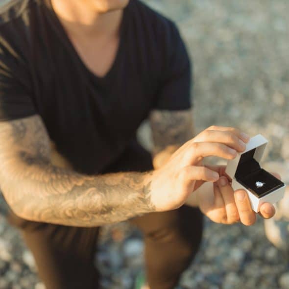 A close-up shot of a man on his knee with tattoos on his arm holding a white box open to reveal an engagement ring.