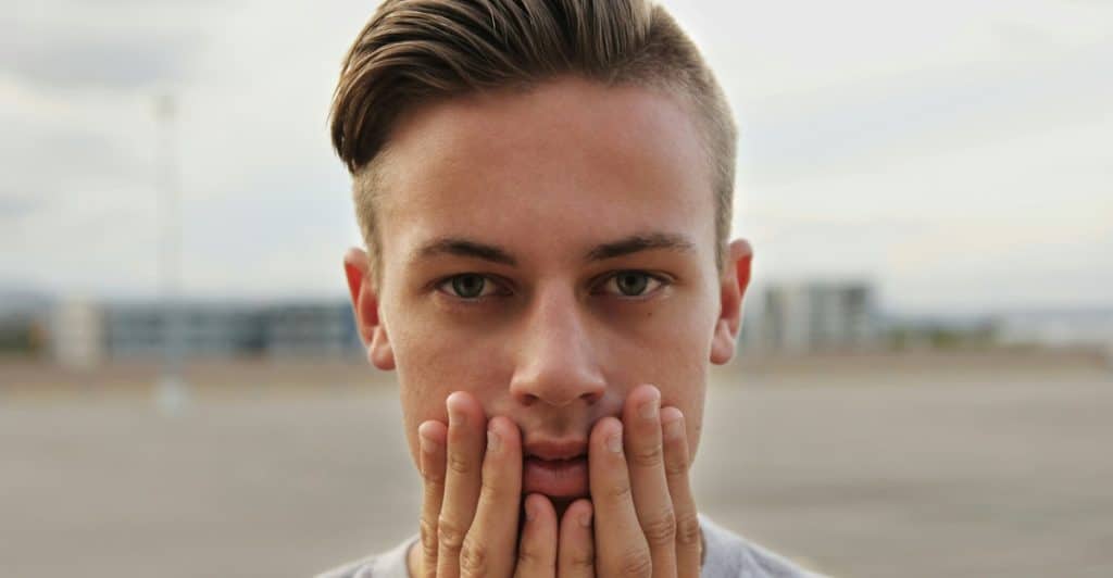 A close-up portrait of a man with light brown hair and blue eyes, wearing a grey t-shirt, with his hands on his face.