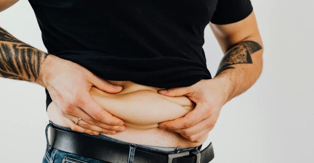 A man in a black t-shirt and jeans holds the skin on his stomach with both hands.