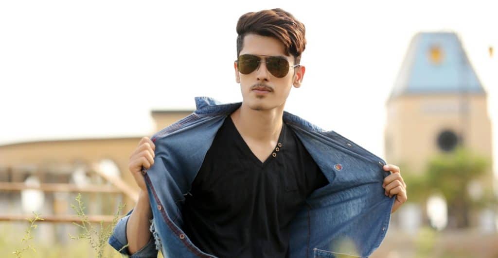 A man with styled hair and sunglasses is holding open a blue button-up shirt, revealing a black v-neck t-shirt.