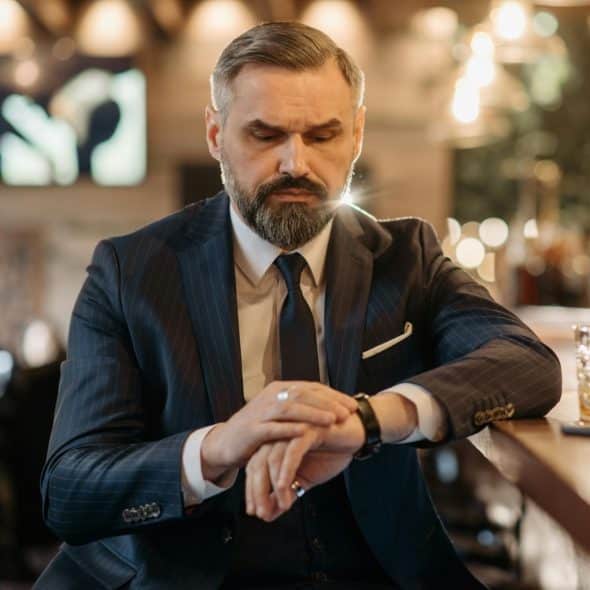 A man in a suit and tie sitting at a bar and looking down at his wrist to check his watch.