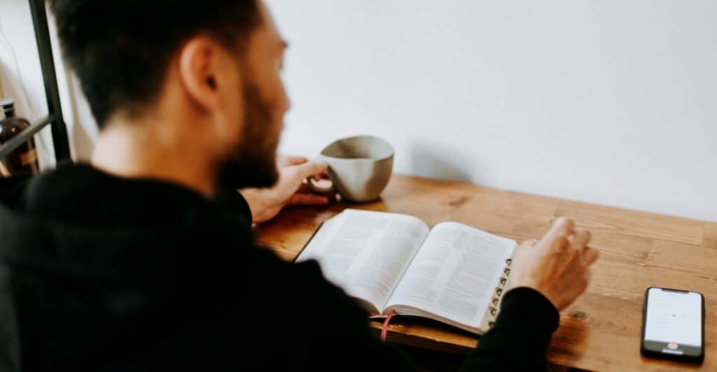 Man wearing a black hoodie sitting at a wooden desk with a book open in front of him and a mug to his left.