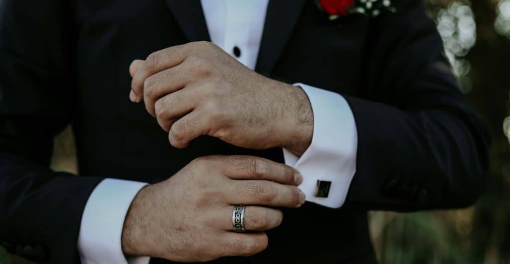 A close-up of a man's hands, wearing a black suit jacket with white cuffs and square cufflinks, adjusting one cuff while wearing a patterned ring on his finger.