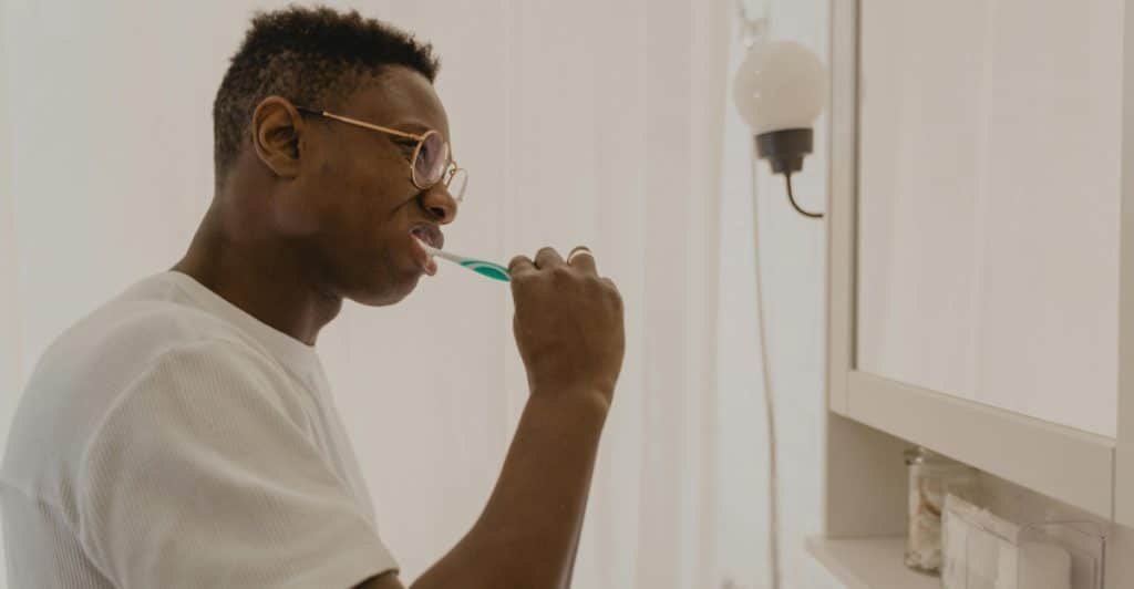 A person in glasses brushing their teeth in a bathroom.