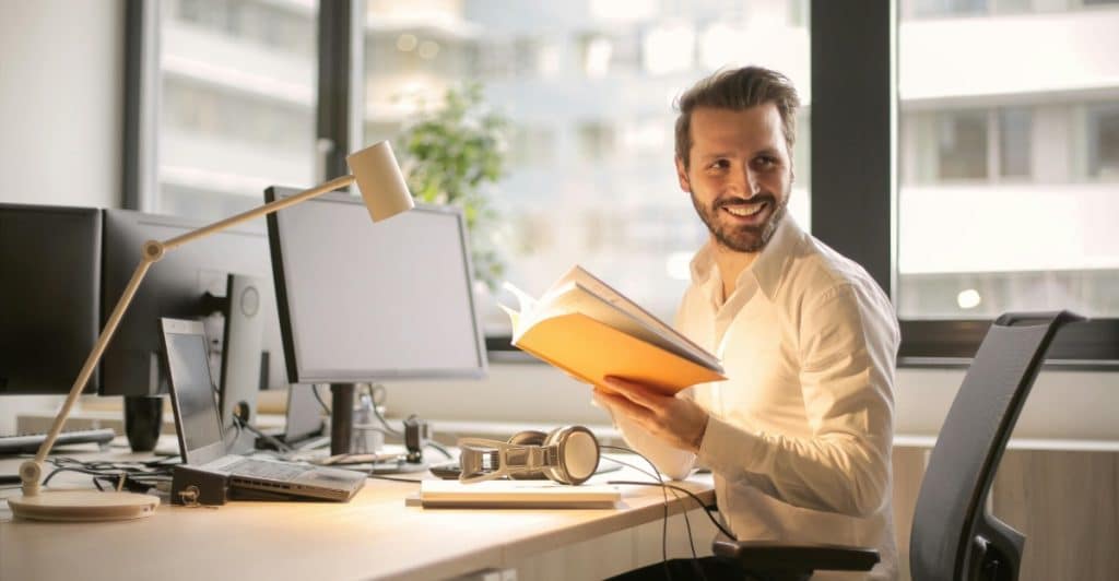 A smiling man with a beard and glasses sitting at a desk with computer monitors and headphones, holding an open book.