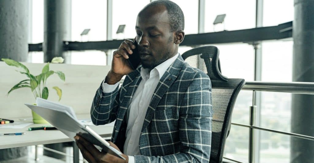 A Black man in a grey and white checkered blazer sits in a chair, holding a document in one hand and talking on a smartphone held to his ear, with a plant and office window visible in the background.