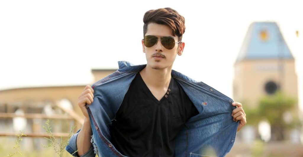 A man in sunglasses and a denim shirt pulling open his shirt to reveal a black t-shirt, with a building in the background.