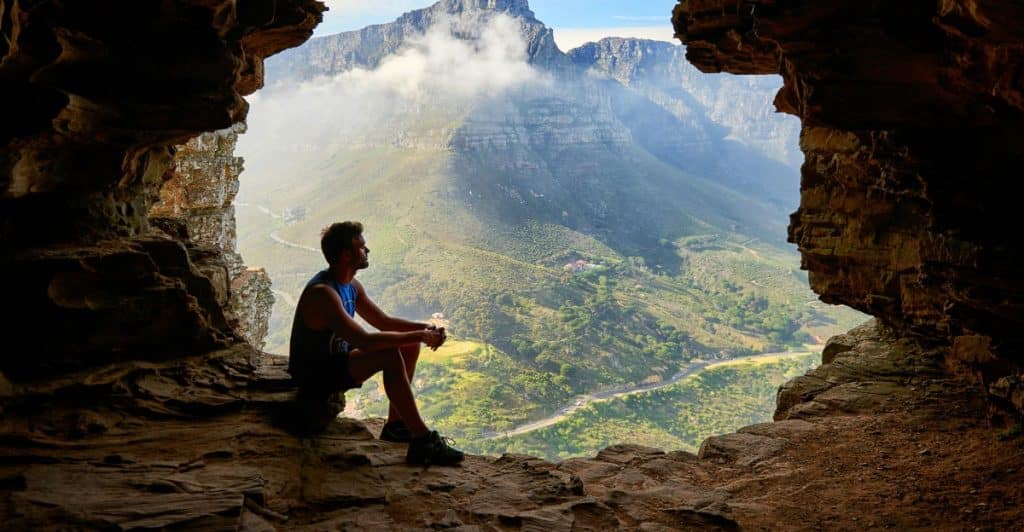 Man sitting at the entrance of a cave with mountains and a valley in the background.