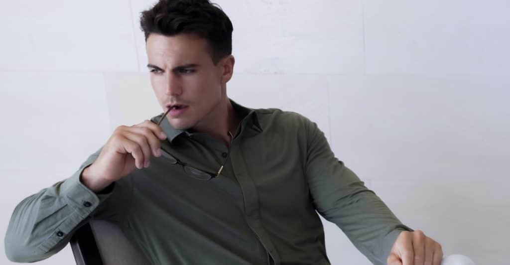 A man in a long-sleeve olive green shirt and white pants, sitting on a chair and holding sunglasses near his face.