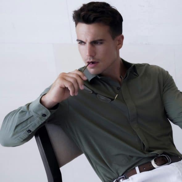 A man in a long-sleeve olive green shirt and white pants, sitting on a chair and holding sunglasses near his face.