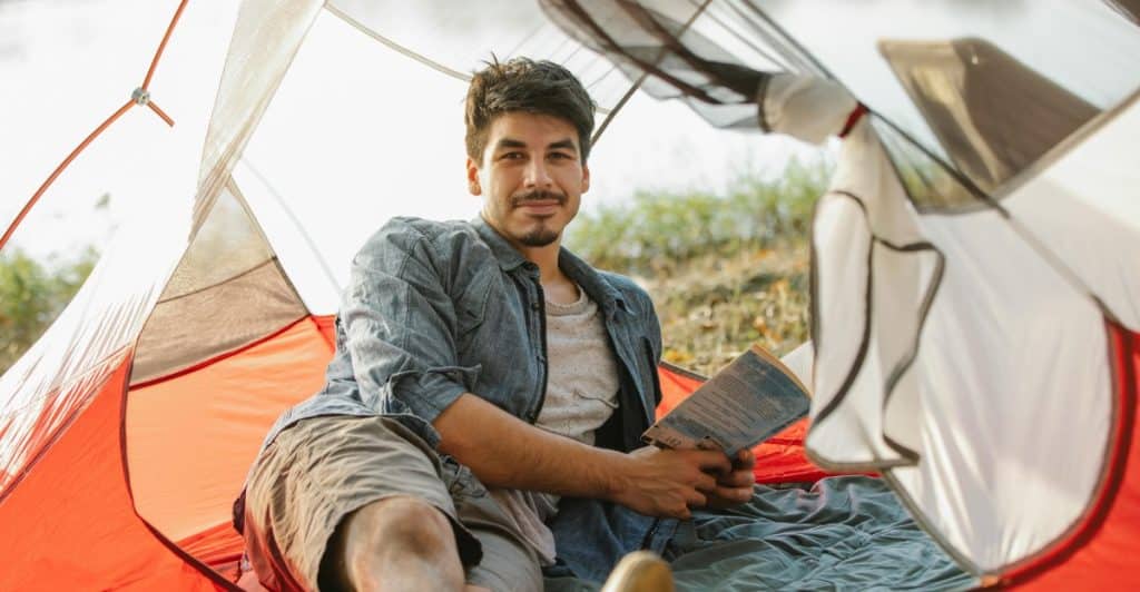A man with dark hair is sitting in a tent, reading a book, and wearing a gray shirt and shorts.