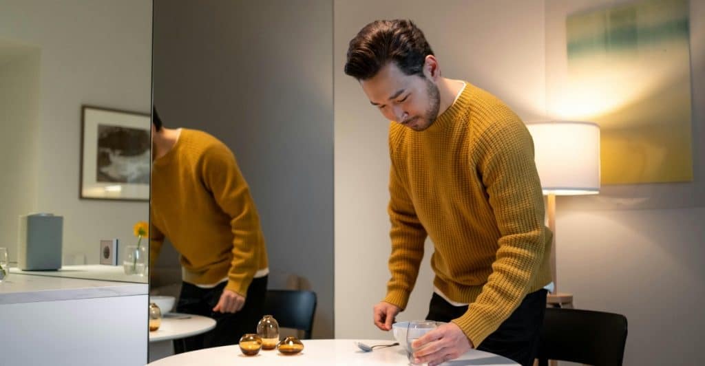 A man wearing a yellow sweater standing in an apartment with a mirror and a table.