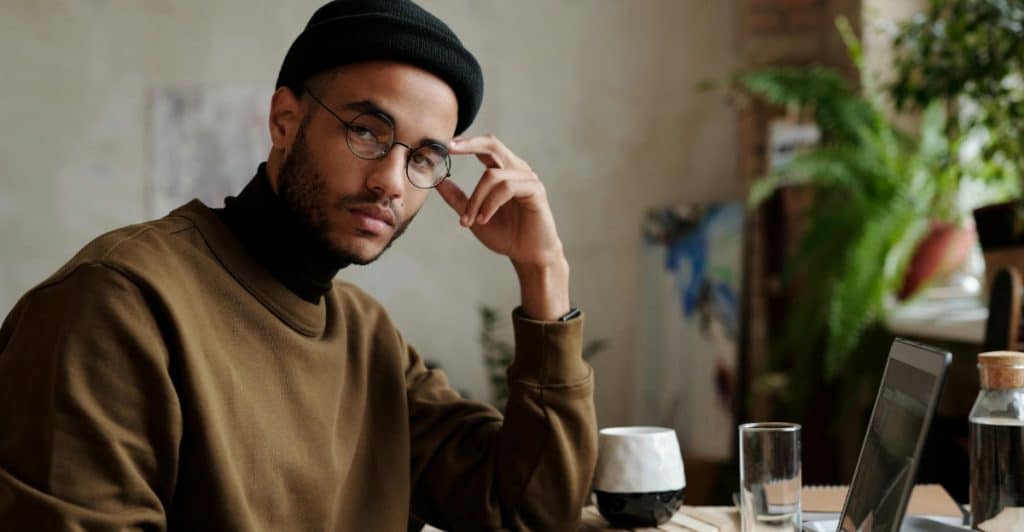 A man wearing a beanie and glasses sits at a table with a laptop.