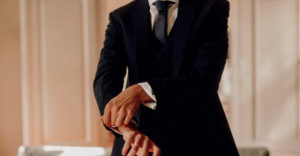 A man in a black suit and tie adjusting his cuff.