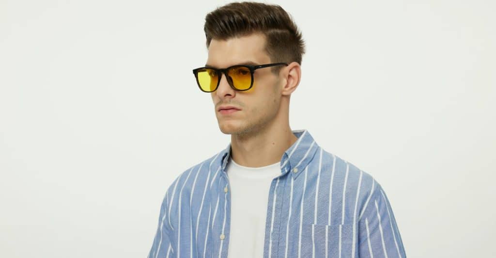 A man with styled hair is wearing yellow-tinted glasses and a blue striped button-down shirt over a white t-shirt.