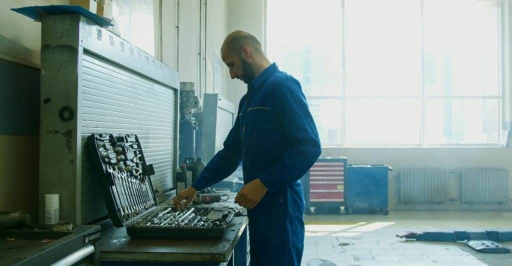 A bald man in a blue jumpsuit reaches into an open toolbox filled with various tools, in a garage or workshop setting.