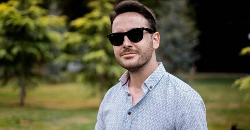 A man with dark hair and sunglasses wearing a light blue patterned button-down shirt and smiling slightly in an outdoor setting.