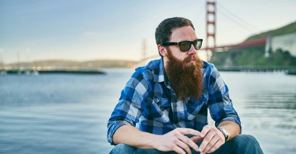 : A man with a beard and sunglasses sitting outdoors, wearing a blue and white plaid shirt.