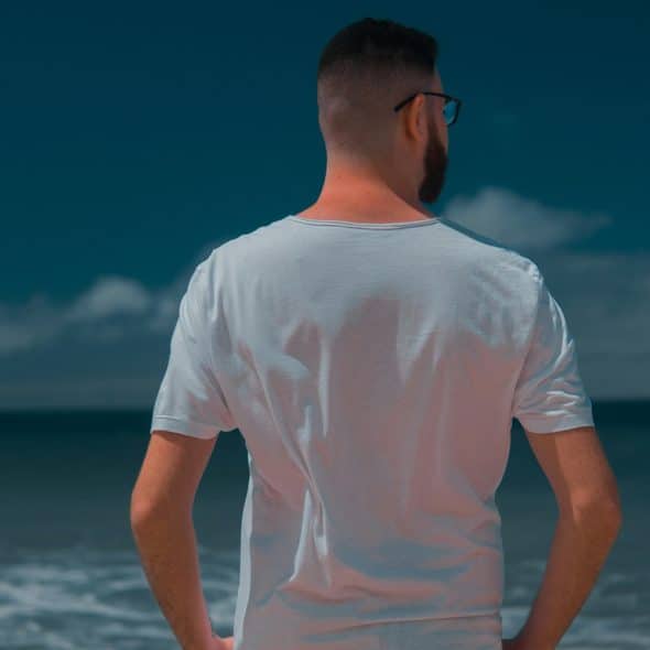 A man with a beard and glasses, wearing a white t-shirt, stands with his back to the viewer,