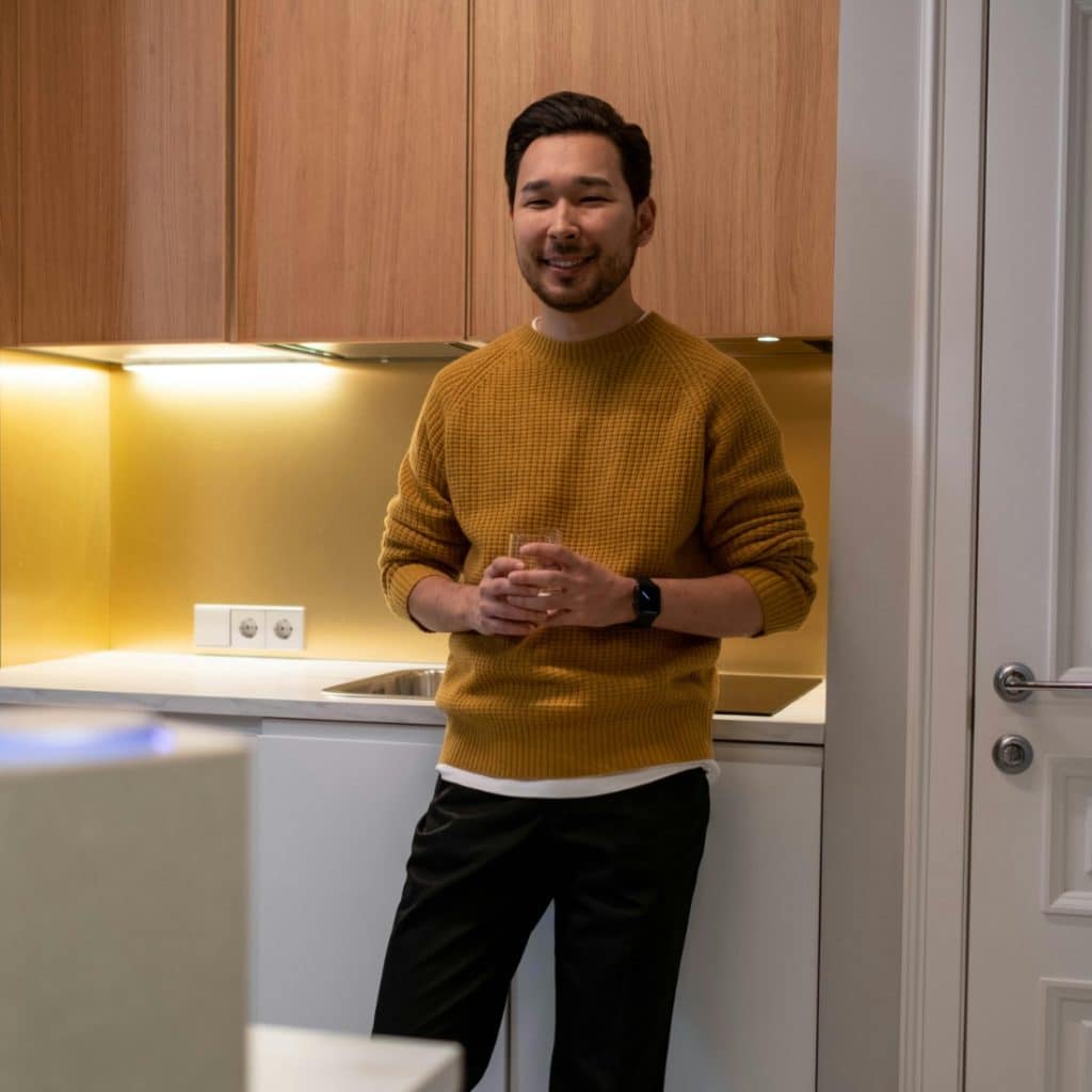 A man wearing a yellow sweater standing in a modern kitchen with light wood cabinets and smiles at the camera.