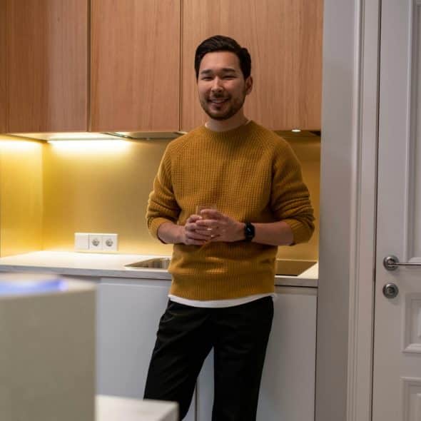 A man wearing a yellow sweater standing in a modern kitchen with light wood cabinets and smiles at the camera.