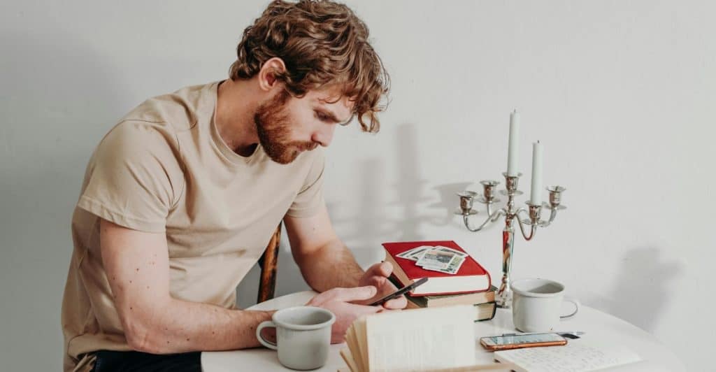 A man with curly hair sitting at a table with books and candles, writing in a journal.