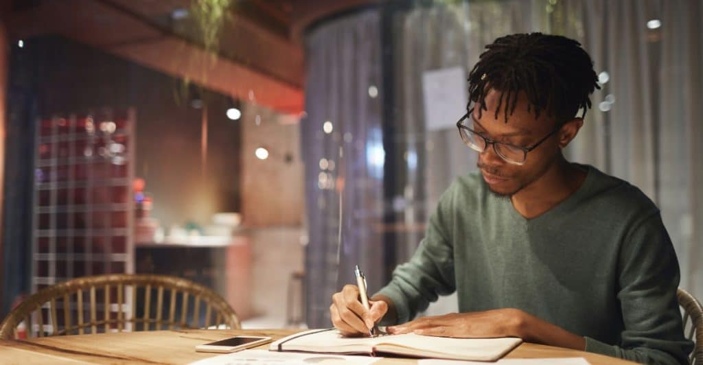  A young dark-skinned man with glasses writing in a notebook at a wooden table in a warm-lit setting.