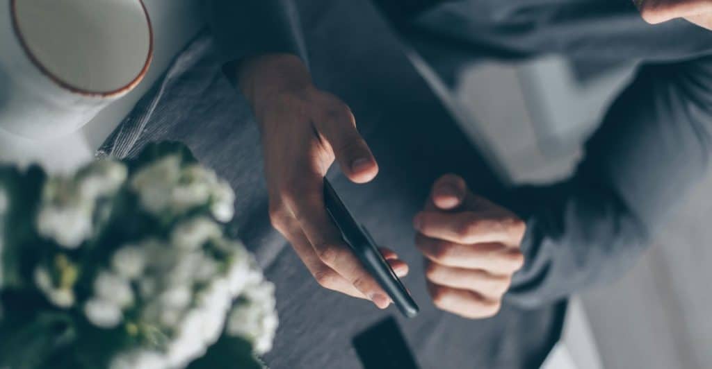  A man looking down at his phone, with papers, a card, and a partial view of a plant on a table.