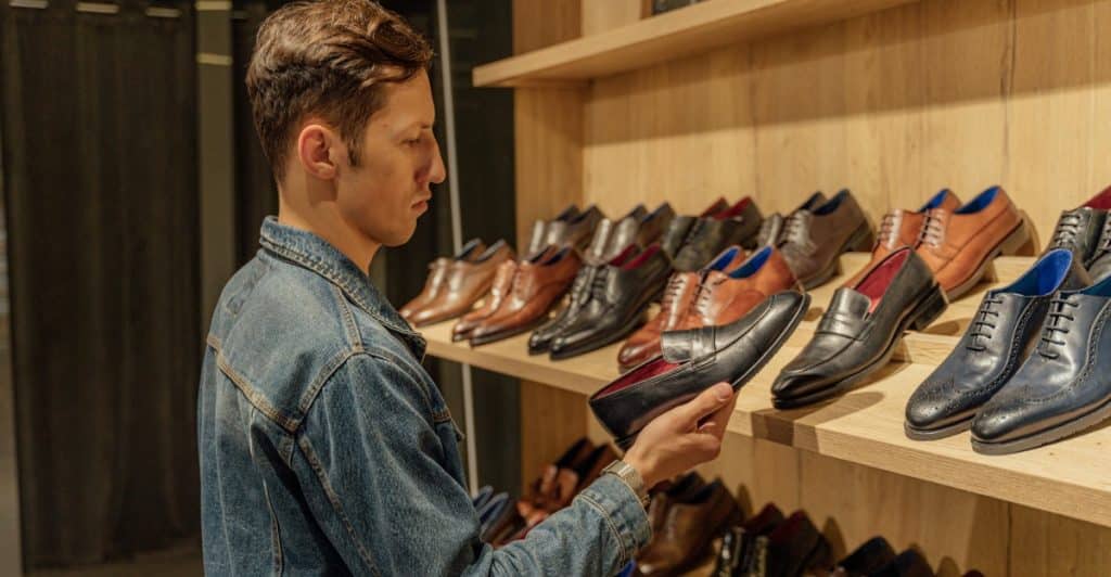 A man in a denim jacket is looking at a selection of shoes on shelves in a store.