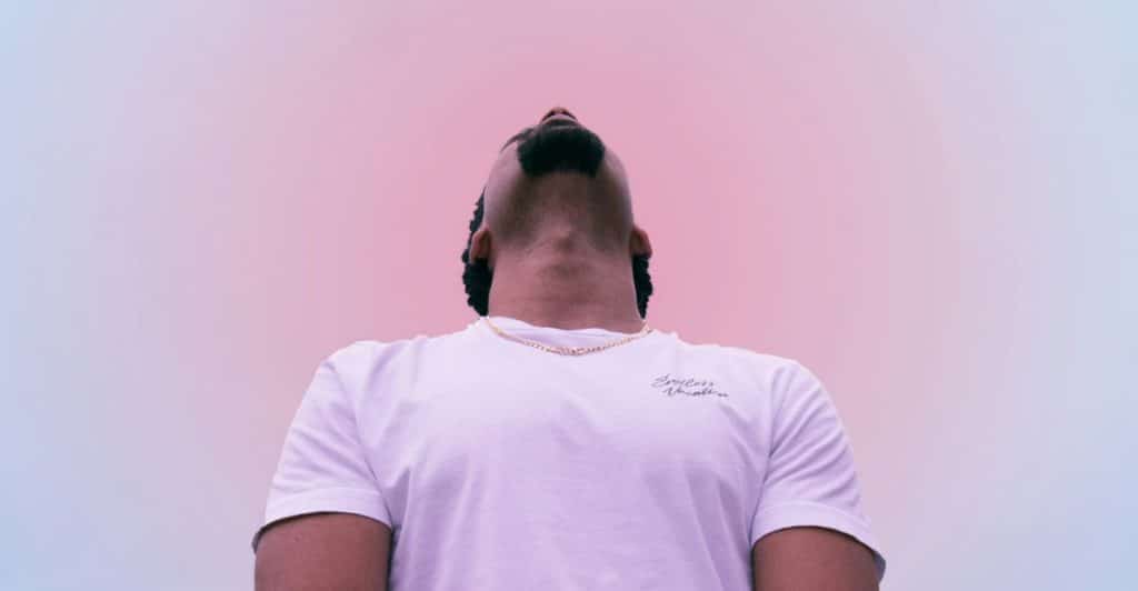 A man with a beard and a white t-shirt looking upwards against a gradient pink and blue background.