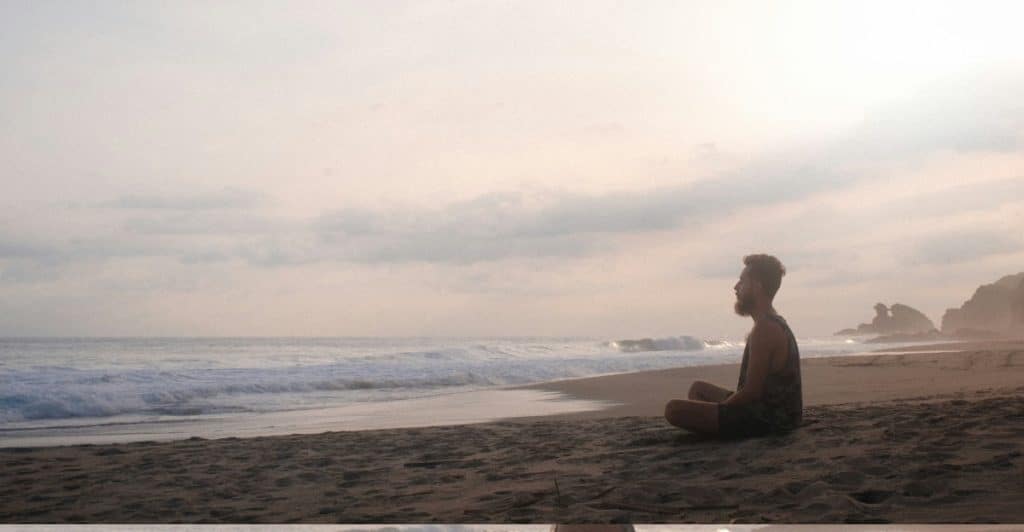 A man is meditating on a beach, sitting in a cross-legged position.