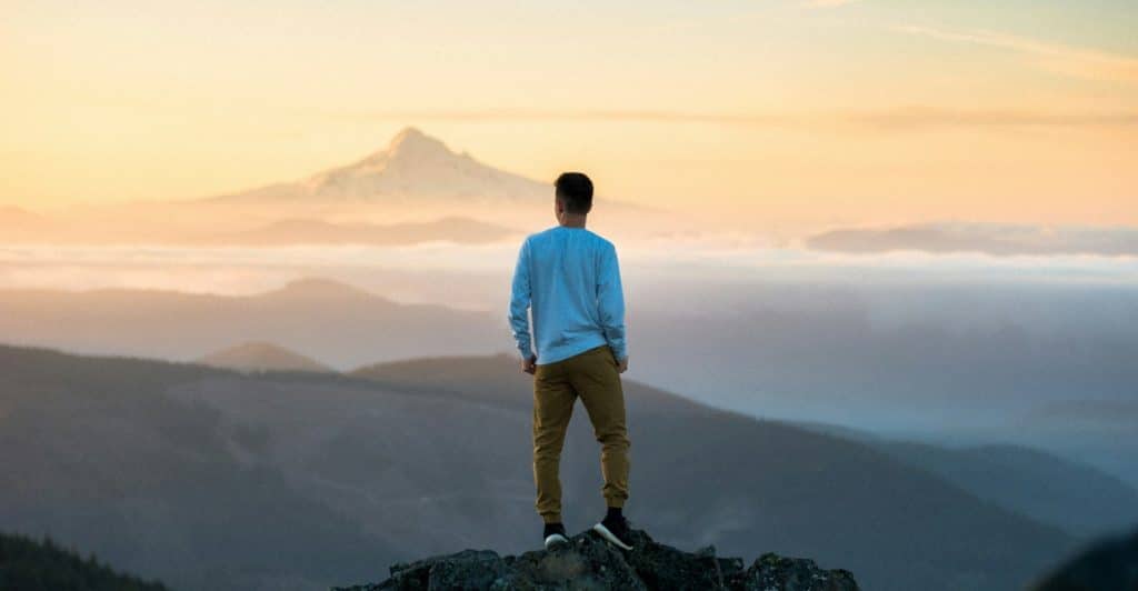 A man standing on a rocky peak, facing away from the camera, and looking out at a vast landscape of mountains and clouds during sunrise.