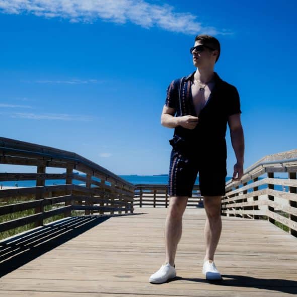 A man wearing sunglasses and a black patterned short-sleeve shirt and shorts, standing on a wooden boardwalk by the beach, looking cool and relaxed.
