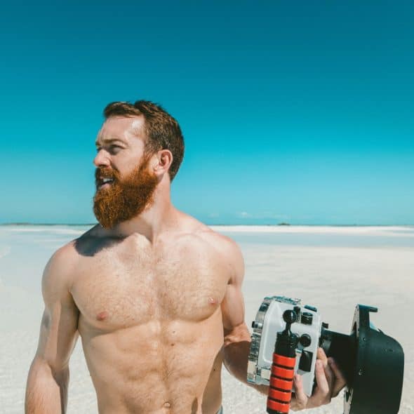 A shirtless man with a beard holds a camera on a bright, white sand beach with a clear blue sky.