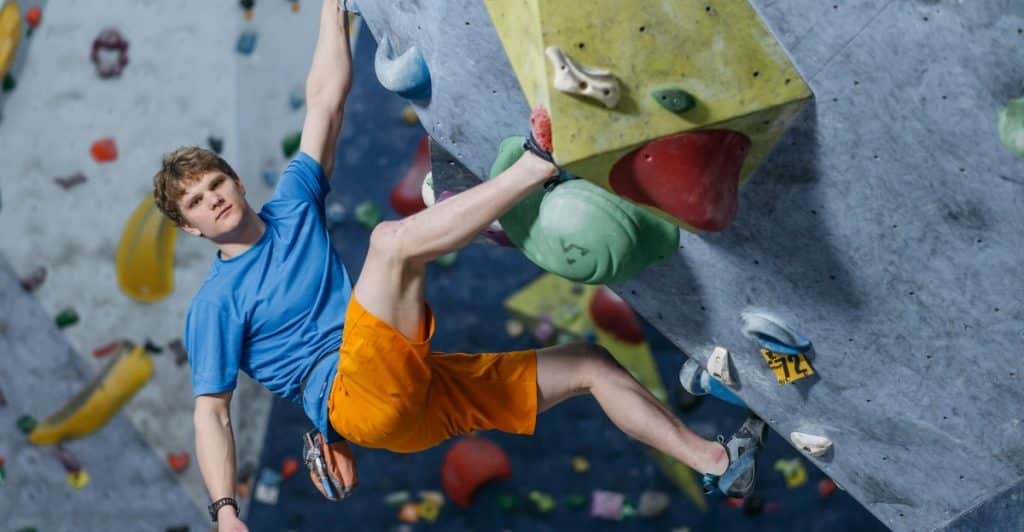 A man wearing a blue t-shirt and orange shorts climbing a colorful indoor climbing wall.